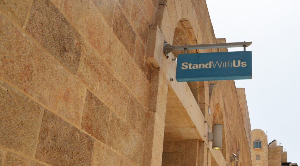 Blue sign reading 'Stand With Us' hanging over a stone alley between sandstone walls with arches at the end of a sunlit street
