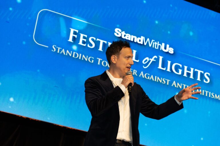 A man in a dark suit speaks into a microphone on stage with a blue event backdrop showing 'Festival of Lights' and 'StandWithUs' text.