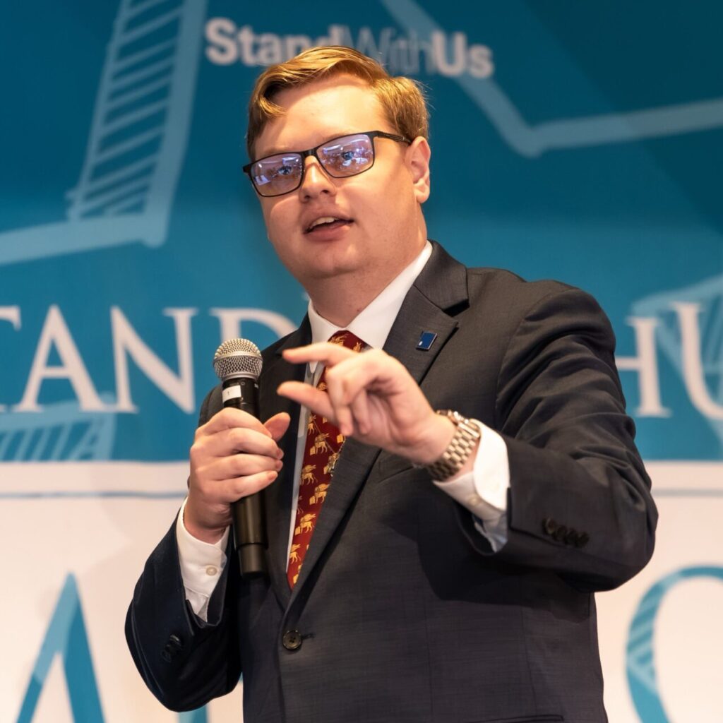 Man in a dark suit and glasses speaks into a microphone on a stage with a blue backdrop reading 'Stand With Us'.