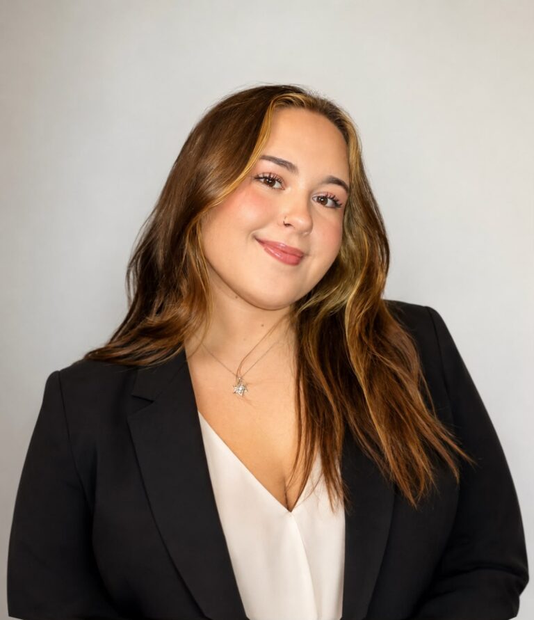 Smiling woman with long brown hair wearing a black blazer and white blouse, posed against a light background.
