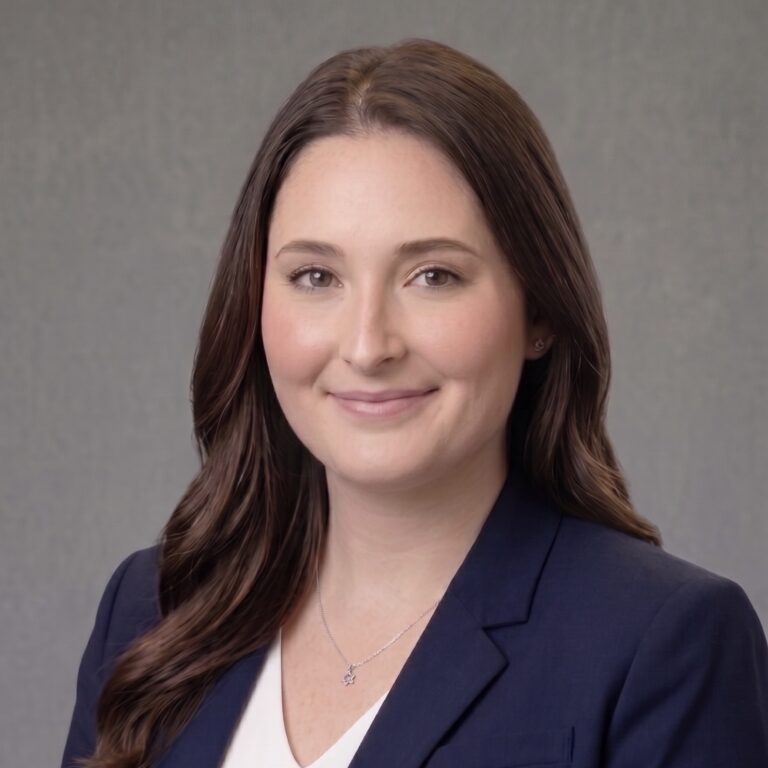 Smiling woman with long brown hair in a navy blazer and white top, posing for a professional portrait against a gray backdrop.
