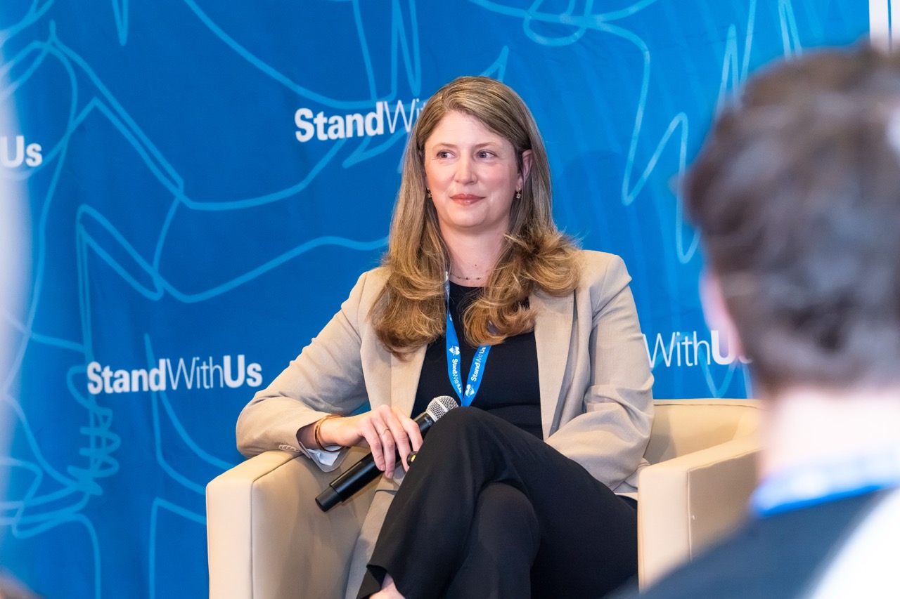 Woman in a beige blazer seated on a chair, holding a microphone, during a panel with a blue StandWithUs backdrop behind her