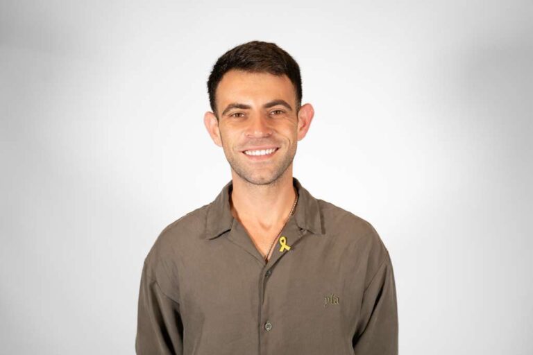 Smiling man in an olive button-down shirt with a yellow awareness ribbon pin, against a light gray gradient background.