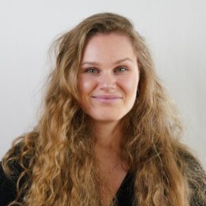 Portrait of a smiling woman with long wavy light brown hair against a pale background, head and shoulders.
