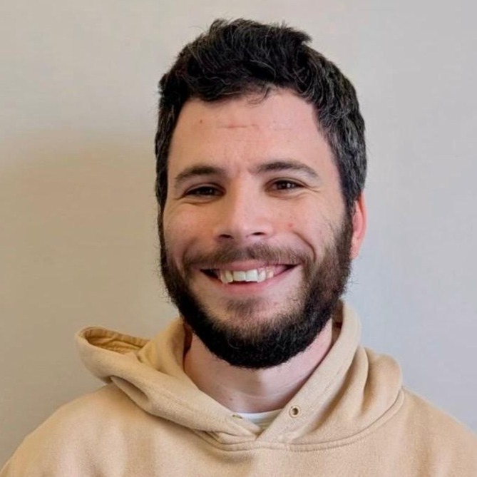 Smiling man with dark hair and beard wearing a beige hoodie with a shaka hand logo on the chest.