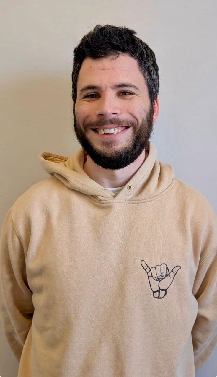 Smiling man with dark hair and beard wearing a beige hoodie with a shaka hand logo on the chest.