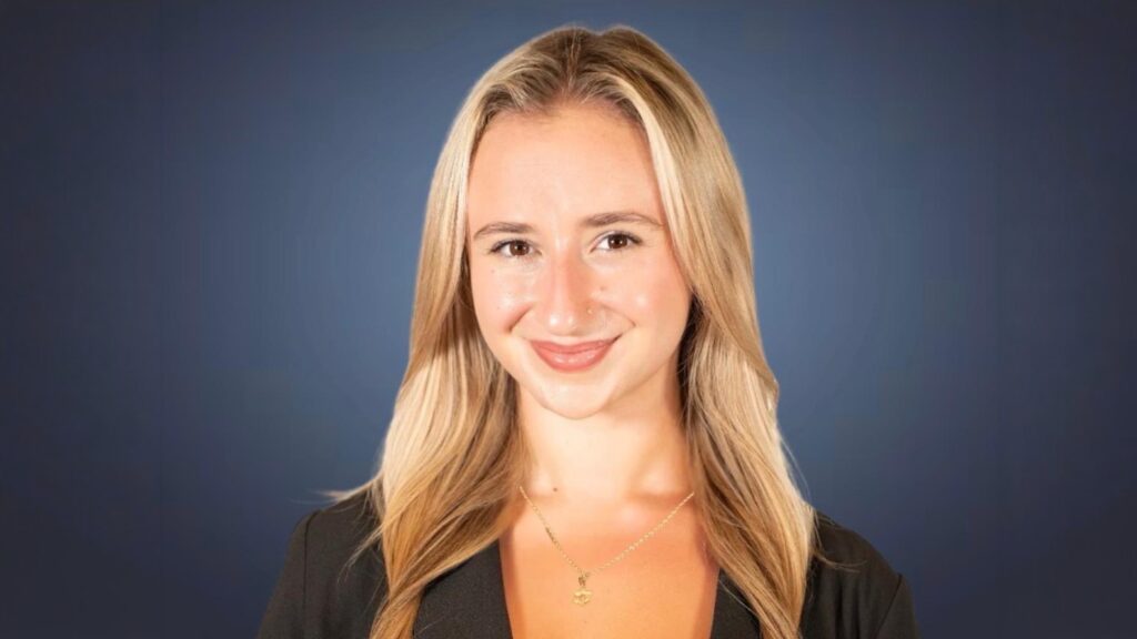 Professional headshot of a smiling blonde woman in a black blazer, against a blue gradient background, wearing a delicate necklace.