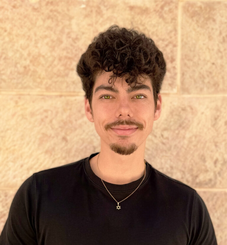 Young man with dark curly hair and a goatee, wearing a black t-shirt, standing against a beige brick wall.