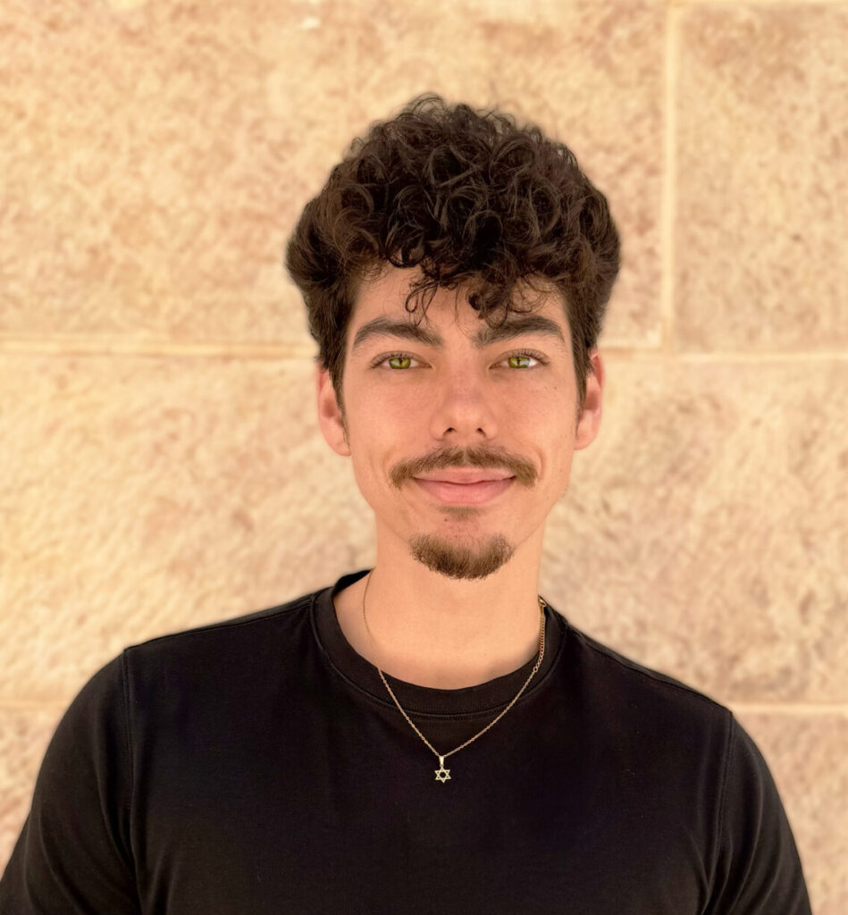 Young man with dark curly hair and a goatee, wearing a black t-shirt, standing against a beige brick wall.