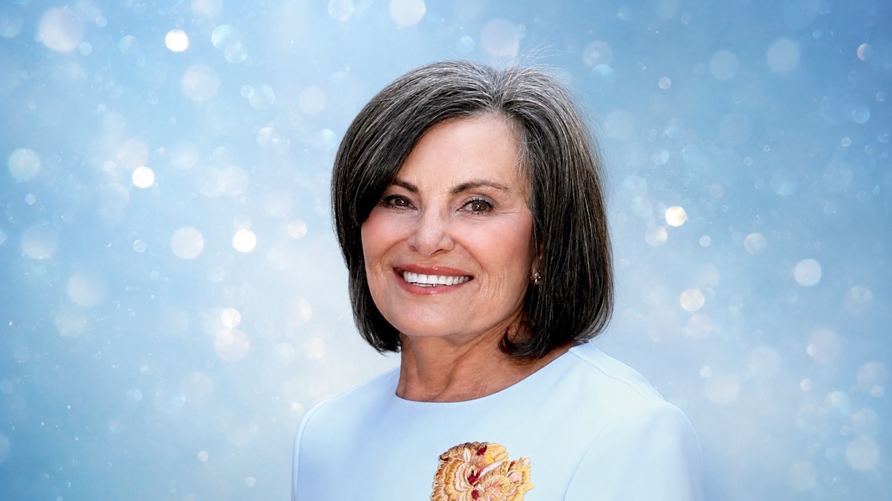 Smiling woman with short gray hair in a light blue dress with a floral brooch against a blue bokeh backdrop.