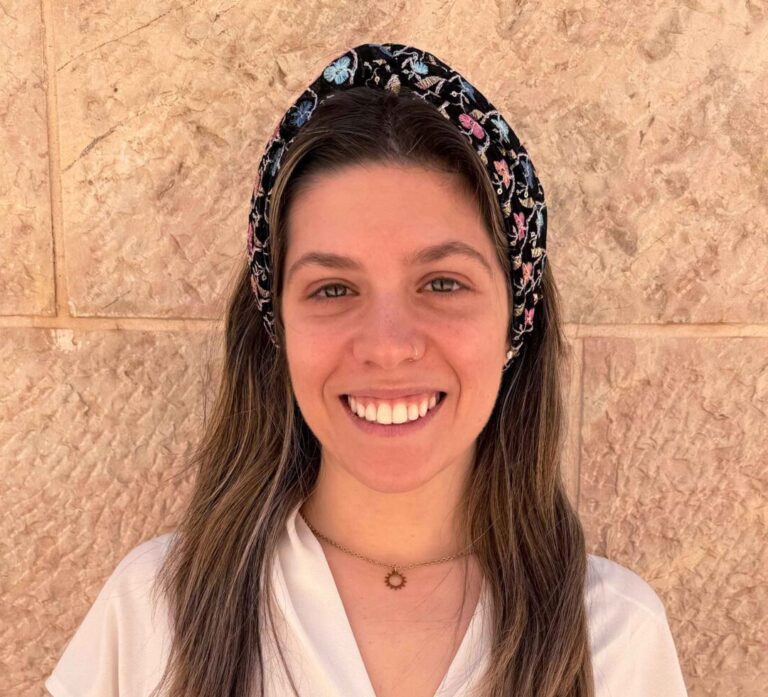 Smiling woman with long brown hair wearing a black floral headband and white blouse, against a beige stone wall.