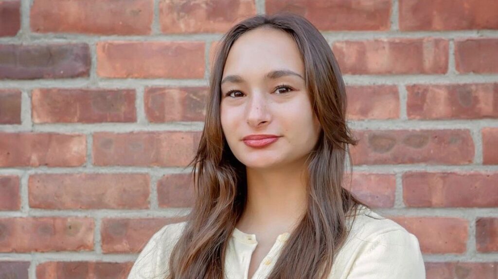 Portrait of a young woman with long brown hair, wearing a light beige top, standing in front of a brick wall.