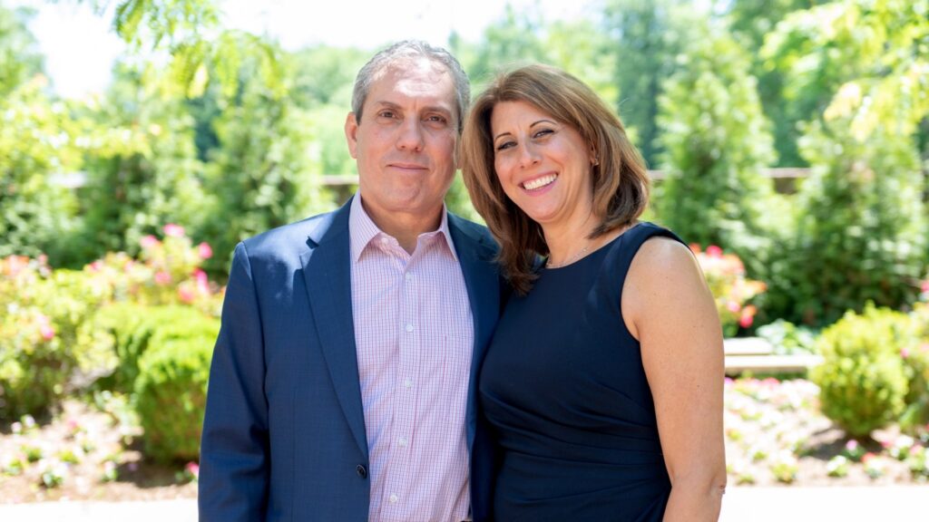 Smiling couple posing together in a sunny garden with greenery and flowers behind them.