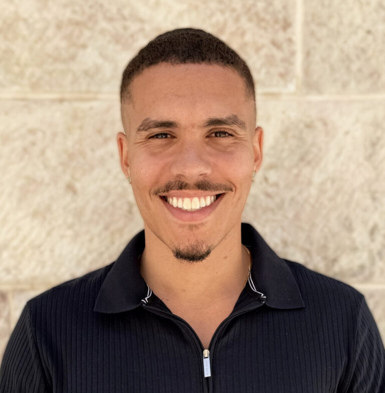 Smiling man with short hair and a light mustache wearing a dark ribbed polo against a beige stone wall.