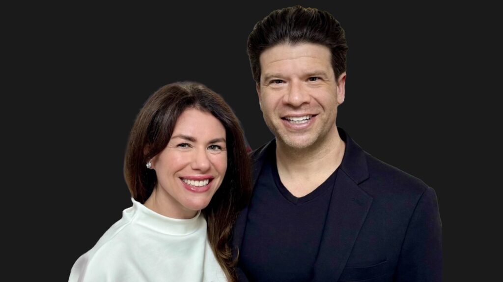 Smiling man and woman posing together against a dark background, shoulders touching.