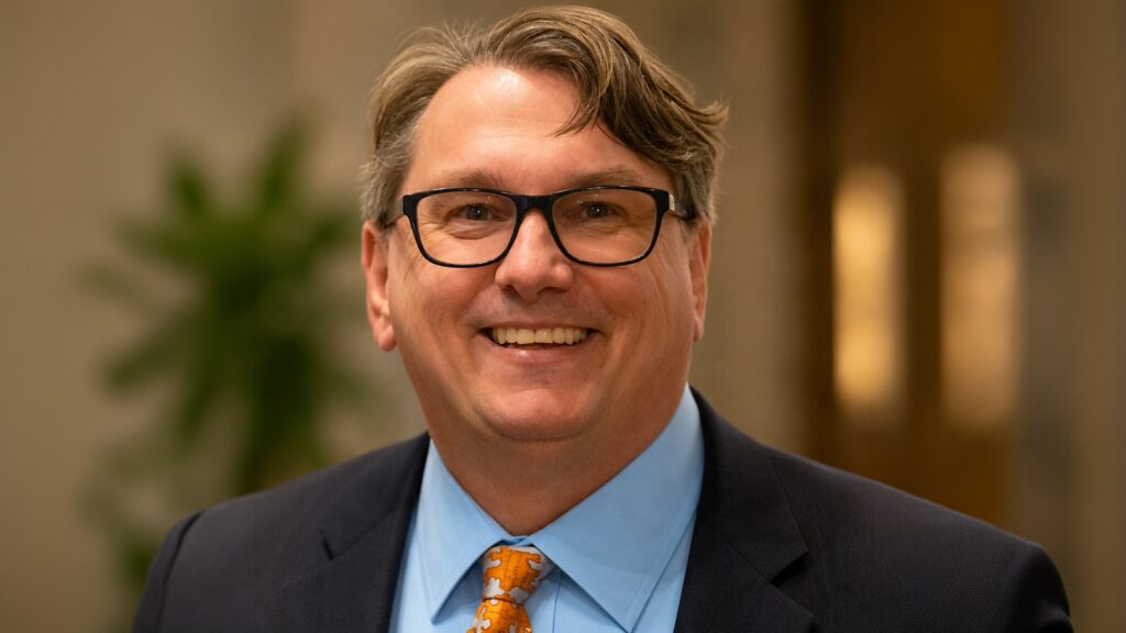 Smiling man in a dark suit with a blue shirt and orange patterned tie, wearing black glasses.
