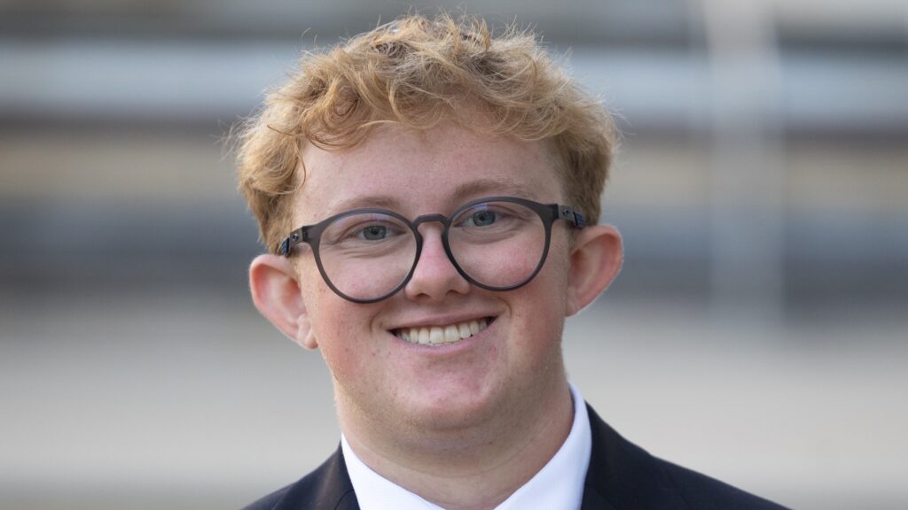 Young man with round black glasses and blond hair smiling outdoors in a suit and tie.