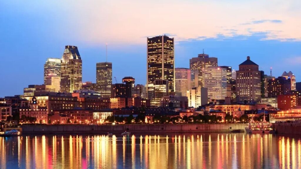 City skyline at dusk reflecting on a calm river, with tall illuminated buildings.
