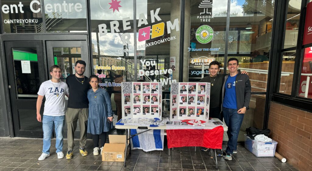 Six people stand outside a storefront with glass windows, next to a table of missing-person posters and a red‑white‑blue flag draped over it.
