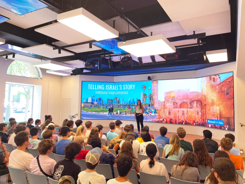 Speaker presenting on stage to a seated student audience, with a large screen behind reading 'TELLING ISRAEL'S STORY.'