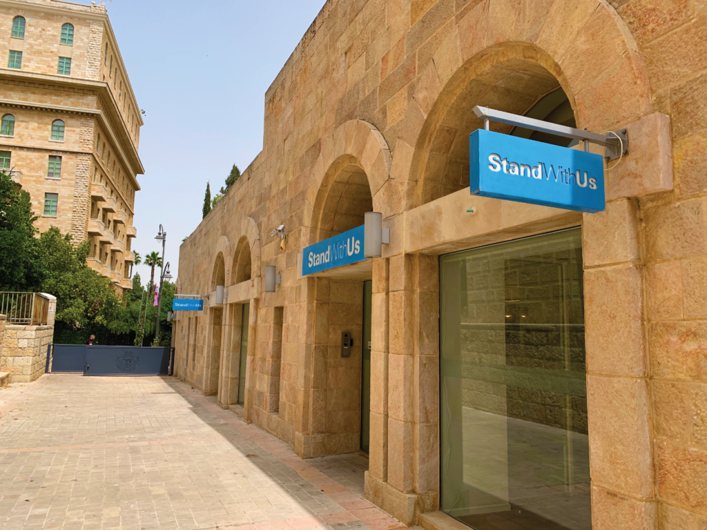 Exterior view of a beige sandstone building with arches and blue StandWithUs signs along a pedestrian path outside the storefronts.”,