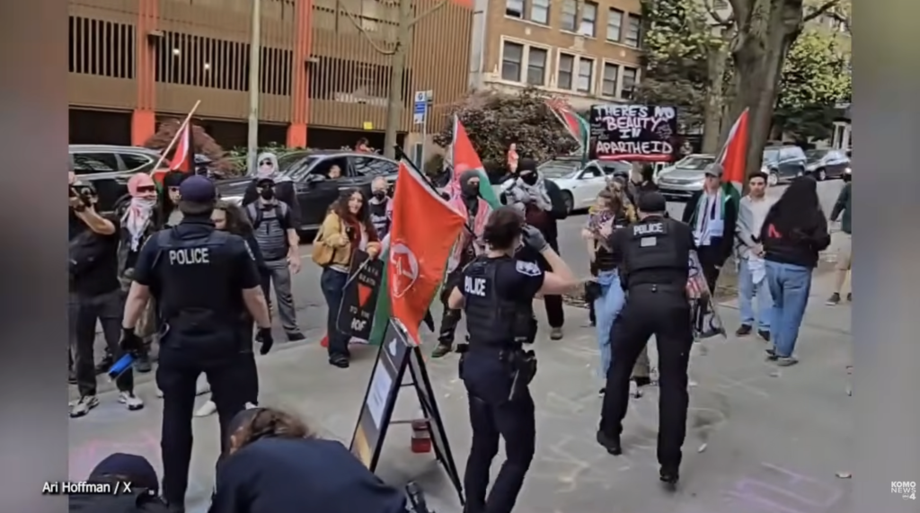 Police in tactical gear face a street protest with Palestinian flags and a banner reading 'There is no beauty in apartheid'.