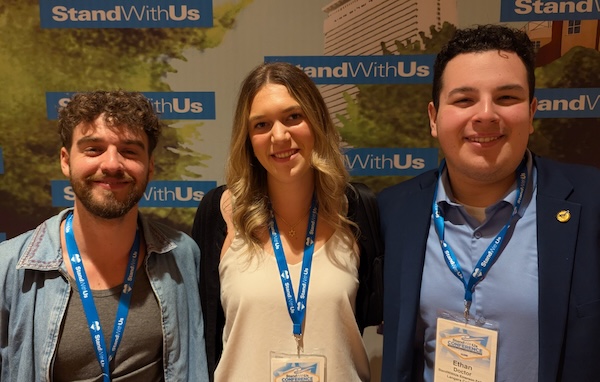 Three smiling attendees wearing blue lanyards pose for a photo at a StandWithUs event with banners in the background.