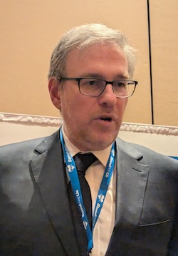 Close-up of a man with gray hair and glasses in a dark suit and tie, wearing a blue conference lanyard, speaking at a formal event.