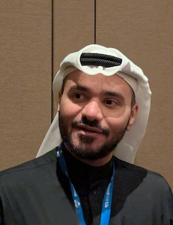 Man in traditional Arabic attire (white ghutra and dark robe) with a blue lanyard, looking to the left.