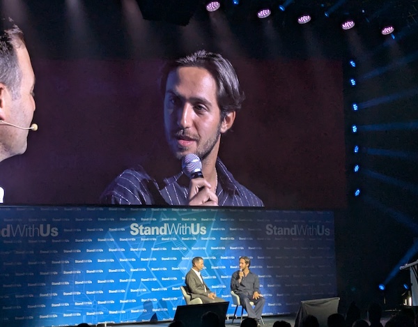 Two men sit on a stage in a talk, one on a large screen holding a microphone. StandWithUs banners line the backdrop below.