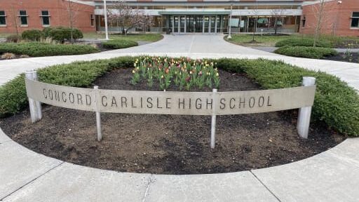Curved concrete sign reads 'Concord Carlisle High School' in front of a flower bed and the school entrance.