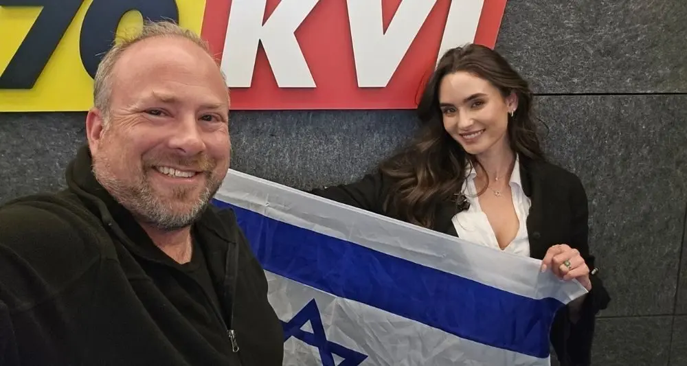 Smiling man and woman pose for a selfie in a broadcasting studio, the woman holding an Israeli flag toward the camera.