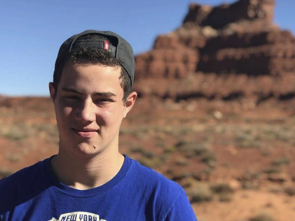 Young man in a blue shirt and backward gray cap standing in a desert with red rock formations in the background.