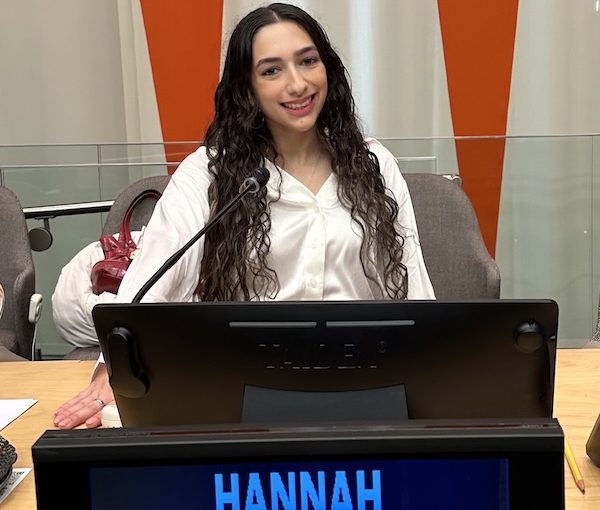 Smiling woman with long dark hair in a white blouse sits at a panel desk with a microphone; name HANNAH appears on the screen.