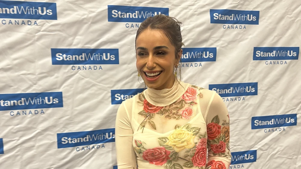download - StandWithUs Smiling woman in a floral sheer top posing in front of a StandWithUs Canada backdrop at a media event.