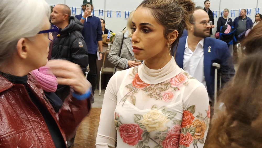 Young woman with a floral turtleneck looks to the side as she talks to an older woman at a crowded indoor event, background attendees visible.