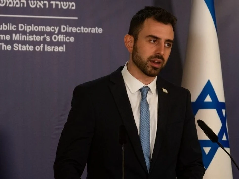 Man in a dark suit and light blue tie speaking at a podium, with an Israeli flag in the background and a backdrop that reads 'Public Diplomacy Directorate' and 'The State of Israel'
