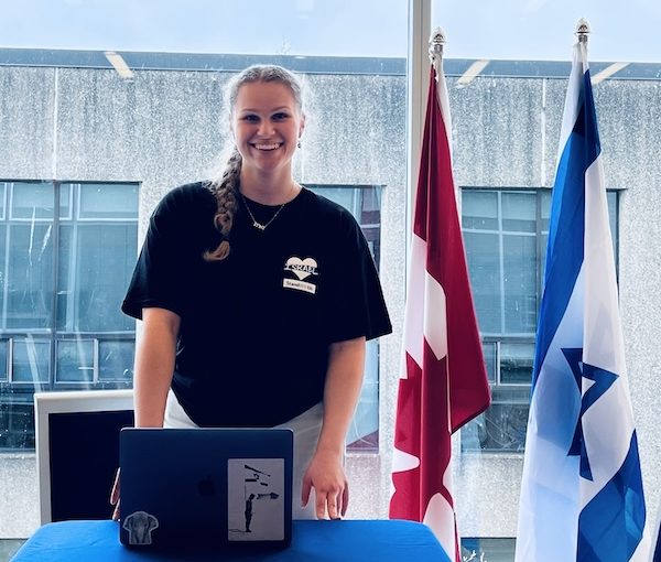 Smiling woman in a black t‑shirt stands behind a blue table with a laptop; Canadian and Israeli flags visible beside her.