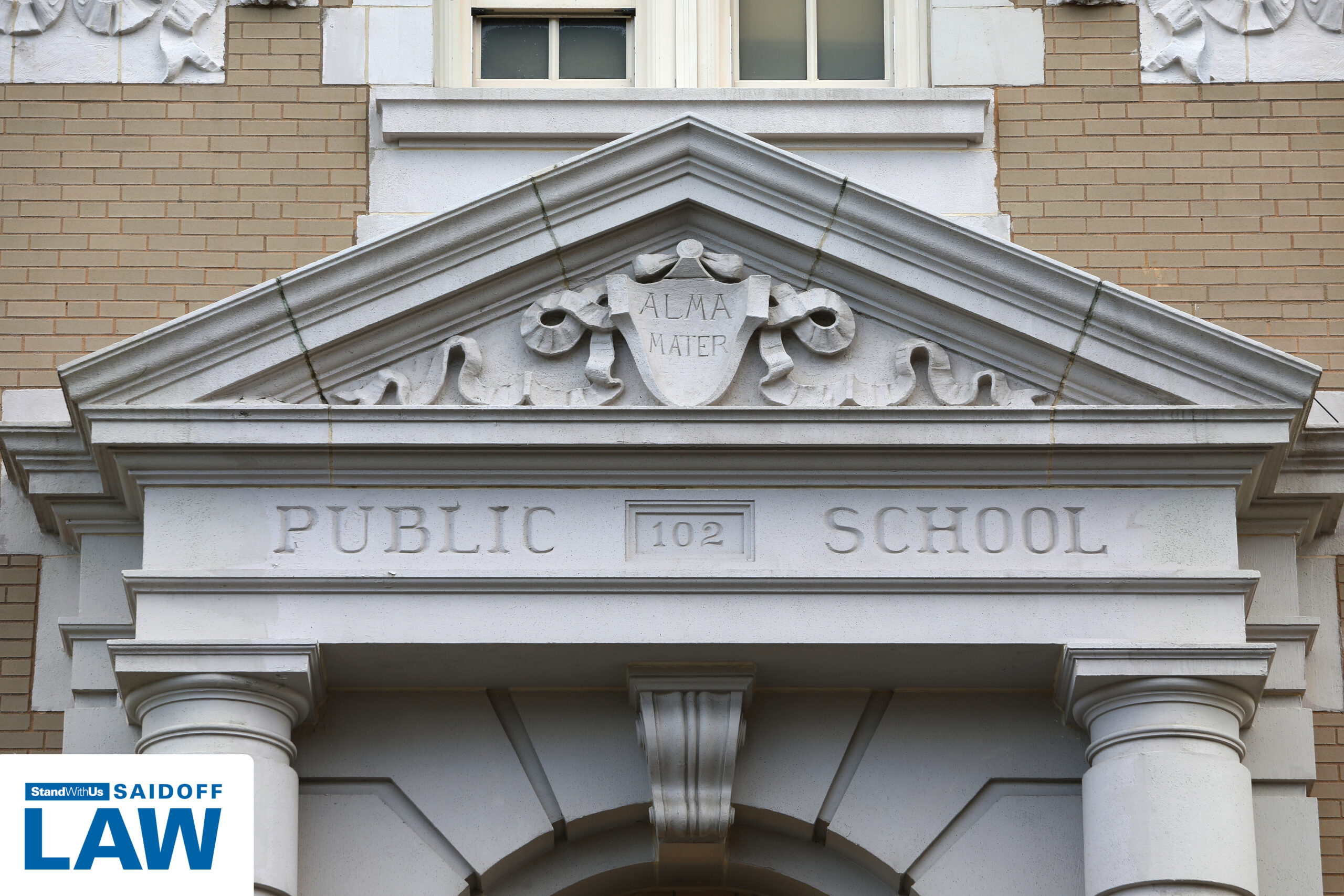 Stone pediment above a public school entrance with palm-shaped scrolls and an ALMA MATER shield crest against a brick wall, and a partial logo in the bottom left corner.