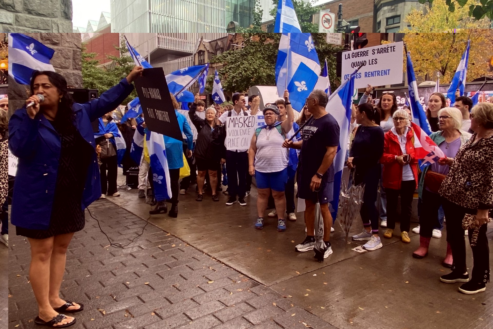Crowd of protesters with blue Quebec flags on a city street; a woman in a blue jacket speaks into a microphone at the front left.