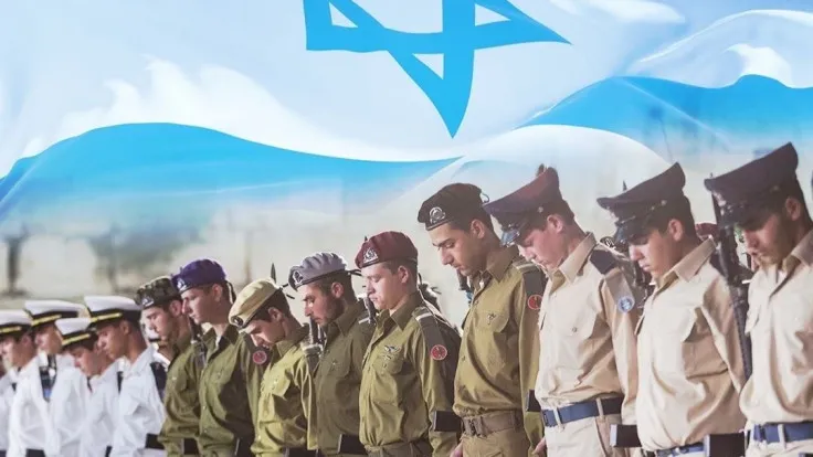 Line of uniformed soldiers bowing heads in a formal ceremony, with a waving Israeli flag in the background.