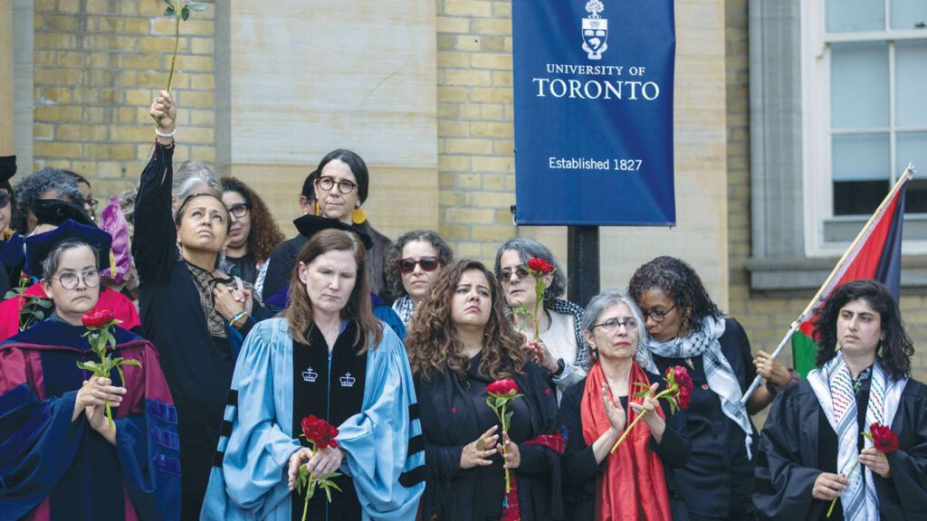 Group of graduates and faculty outdoors in academic robes, some holding red roses, with a University of Toronto banner in the background.