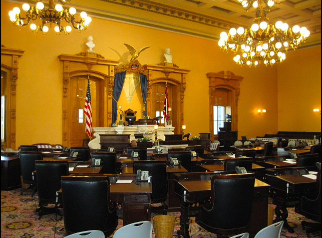 Formal legislative chamber with wooden desks, black chairs, two flags, and an ornate dais with an eagle sculpture and blue drapery.