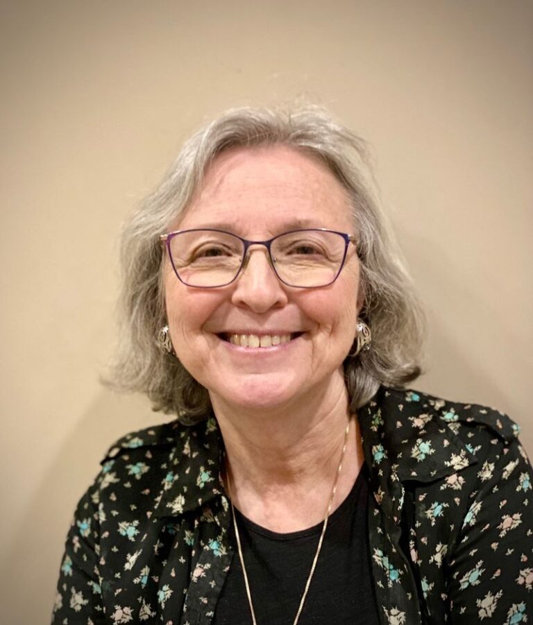 Portrait of a smiling older woman with gray hair and purple-framed glasses, wearing a black floral blouse.