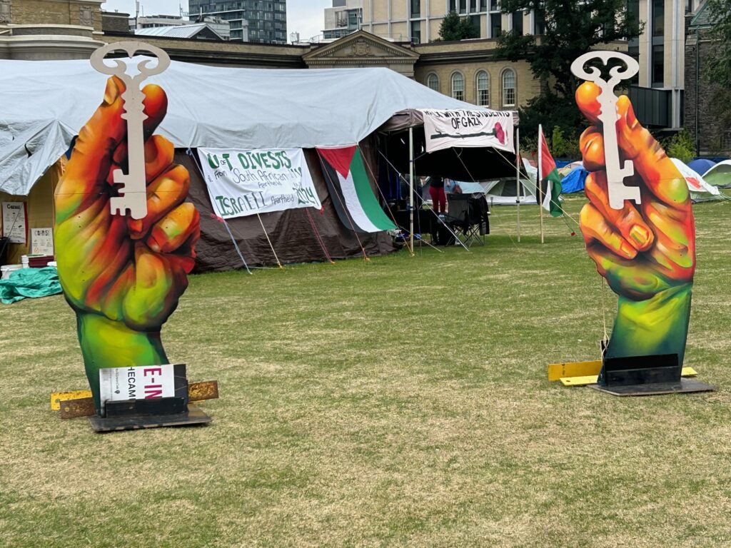 Giant colorful hands holding large white keys stand at a protest camp with tents, Palestinian flags, and a Gaza banner on a grassy area.