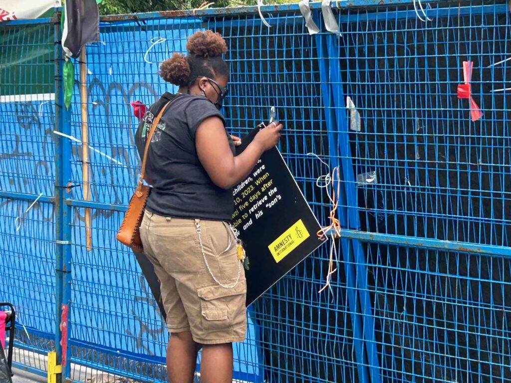 Person with braided hair and a mask stands by a blue metal fence, holding a black protest sign with Amnesty International branding.
