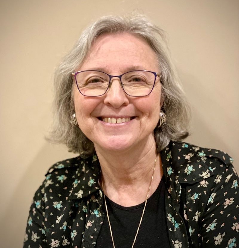 Smiling older woman with gray hair and glasses, wearing a floral black blouse and gold necklace.