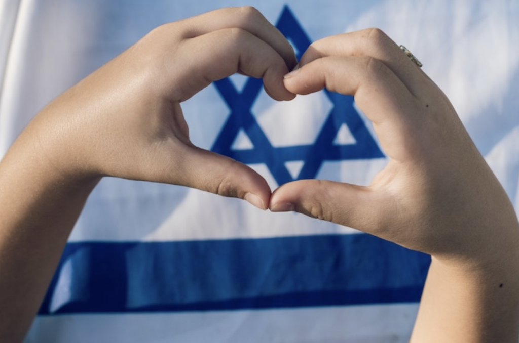 Close-up of hands forming a heart shape in front of an Israeli flag.