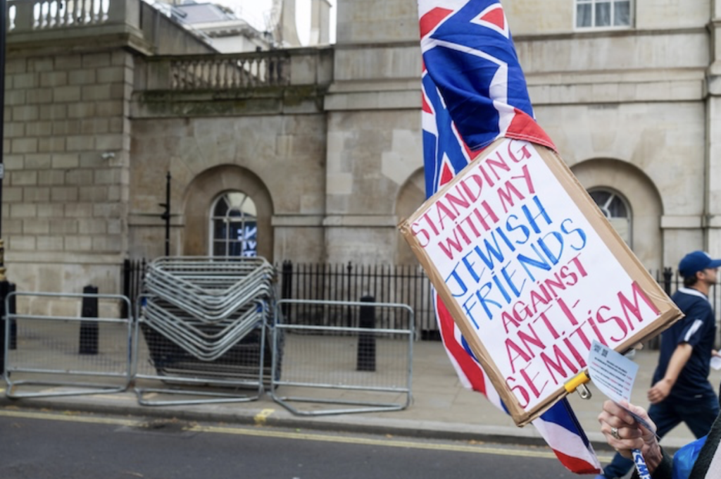 Person holding a protest sign that says 'Standing with my Jewish friends against antisemitism' beside a Union Jack flag on a city street.