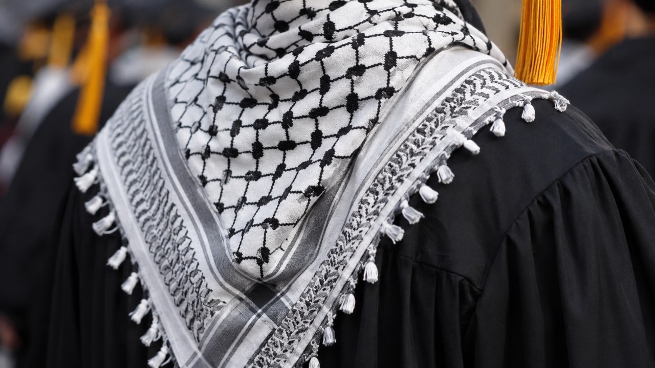 Close-up of a graduate in a black cap and gown with a black-and-white keffiyeh scarf and yellow tassel draped over the shoulder, at a commencement ceremony